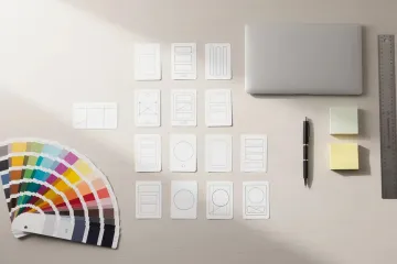 Colour swatches and wireframe layout cards arranged on a desk beside a laptop, pen, sticky notes and ruler